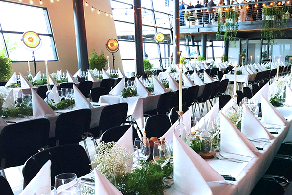 Elegant dukade långbord med vita dukar, vikta servetter, levande ljus och grön växtdekoration i en ljus och rymlig lokal med stora fönster. Lokalen är förberedd för en bankett eller festmiddag, och gäster syns på en övervåning i bakgrunden.