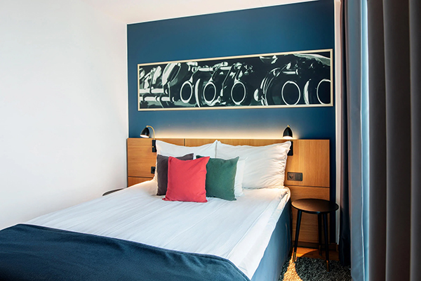 Hotellrum med en fint bäddad säng. Kulörta kuddar på sängen. Mörkblå fondvägg bakom med en svartvit tavla.