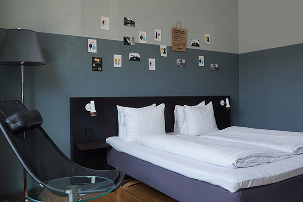 Hotellrum med en fint bäddad säng. Kulörta kuddar på sängen. Mörkblå fondvägg bakom med en svartvit tavla.