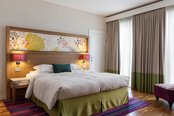 Hotellrum med en fint bäddad säng. Kulörta kuddar på sängen. Mörkblå fondvägg bakom med en svartvit tavla.