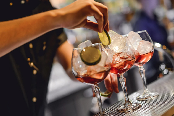 En bartender dekorerar tre cocktails i stora vinglas med is och limeklyftor på en bardisk. Dryckerna har en rödaktig färg och serveras i en livlig bar- eller restaurangmiljö.