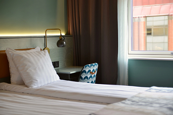 Hotellrum med en fint bäddad säng. Kulörta kuddar på sängen. Mörkblå fondvägg bakom med en svartvit tavla.