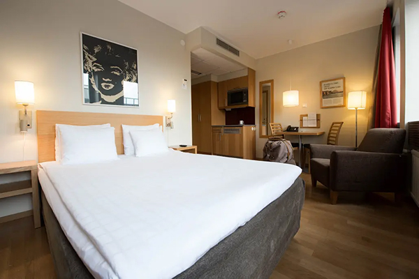 Hotellrum med en fint bäddad säng. Kulörta kuddar på sängen. Mörkblå fondvägg bakom med en svartvit tavla.