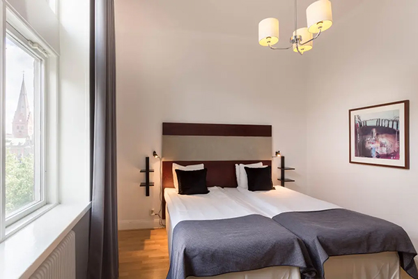 Hotellrum med en fint bäddad säng. Kulörta kuddar på sängen. Mörkblå fondvägg bakom med en svartvit tavla.