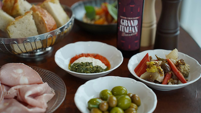 En bartender dekorerar tre cocktails i stora vinglas med is och limeklyftor på en bardisk. Dryckerna har en rödaktig färg och serveras i en livlig bar- eller restaurangmiljö.